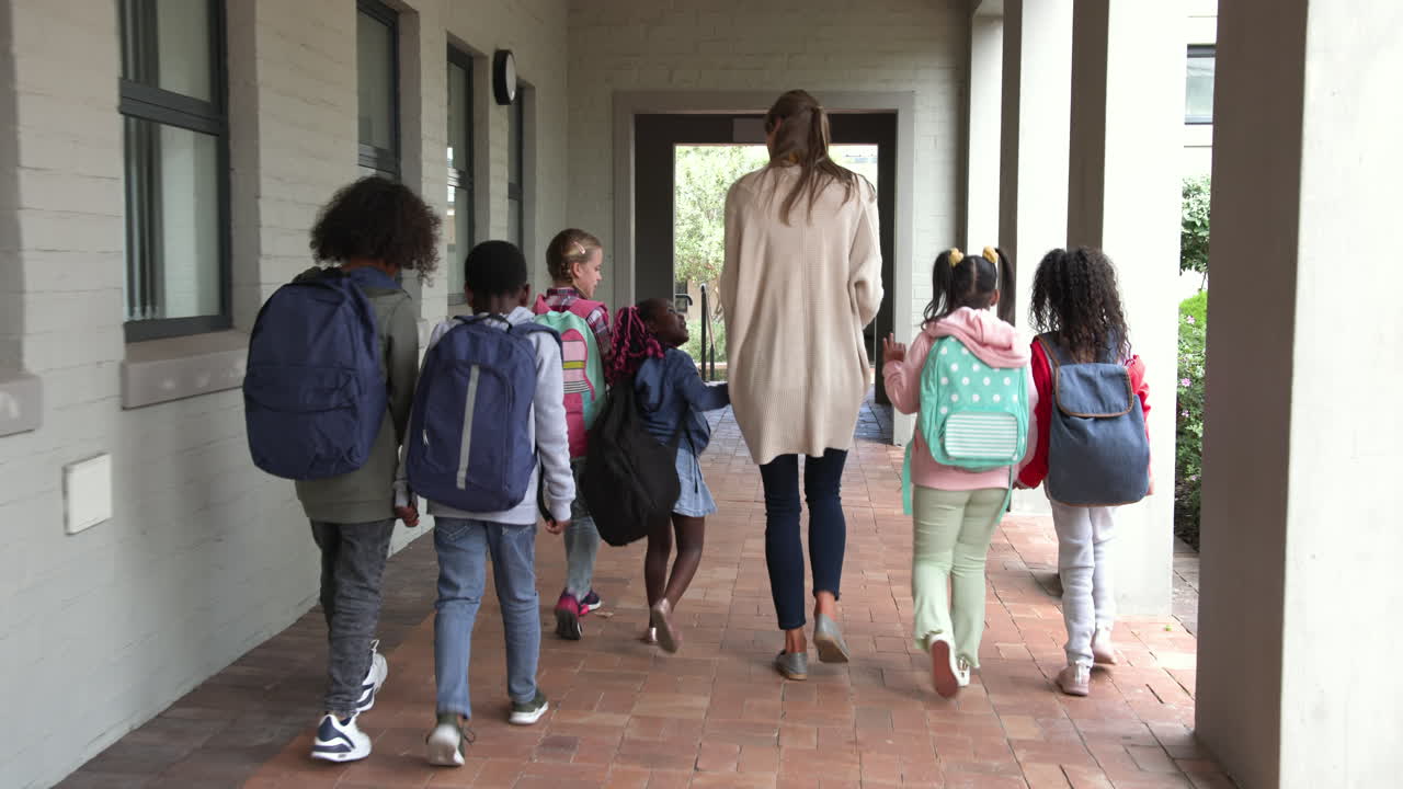 Teacher walking with diverse group of children in school hallway, carrying backpacks
