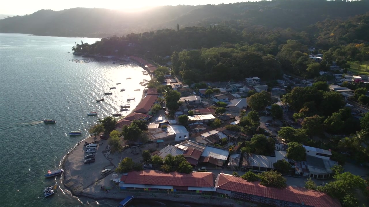 Drone aereo al tramonto che si muove dal lago verso la riva, rivelando barche, case e una grande montagna verde. Paesaggio tropicale sereno ideale per viaggi, turismo e immagini naturalistiche.