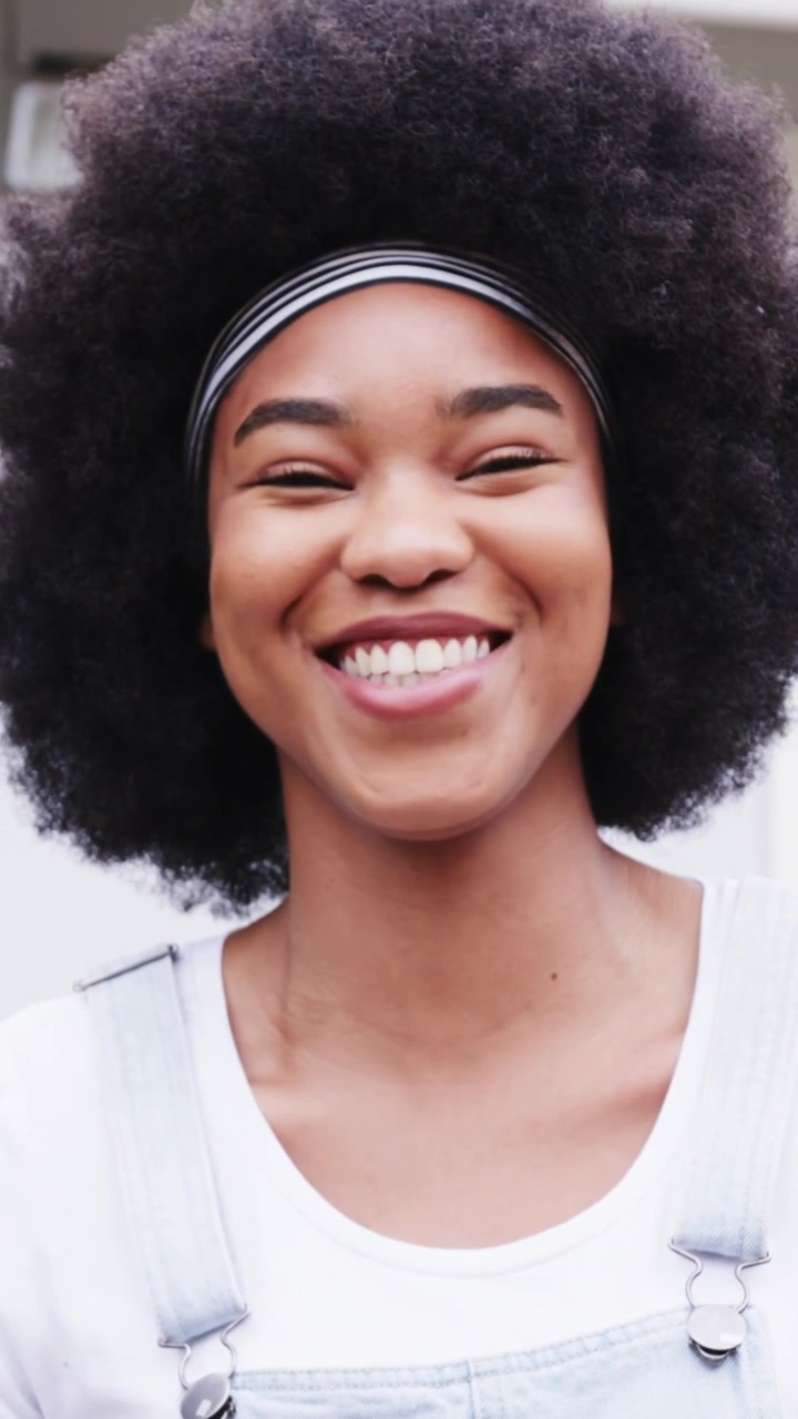 Vídeo vertical: Câmera filmando mulher sorrindo e rindo na porta, tiara listrada e macacão jeans
