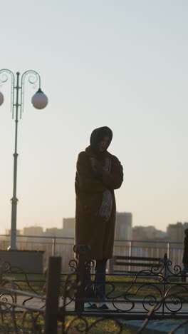 a tearful woman wearing a coat and a scarf stands in front of a stone monument in a park during sunset. moment of sorrow and contemplation, with the warm glow of the setting sun casting long shadows