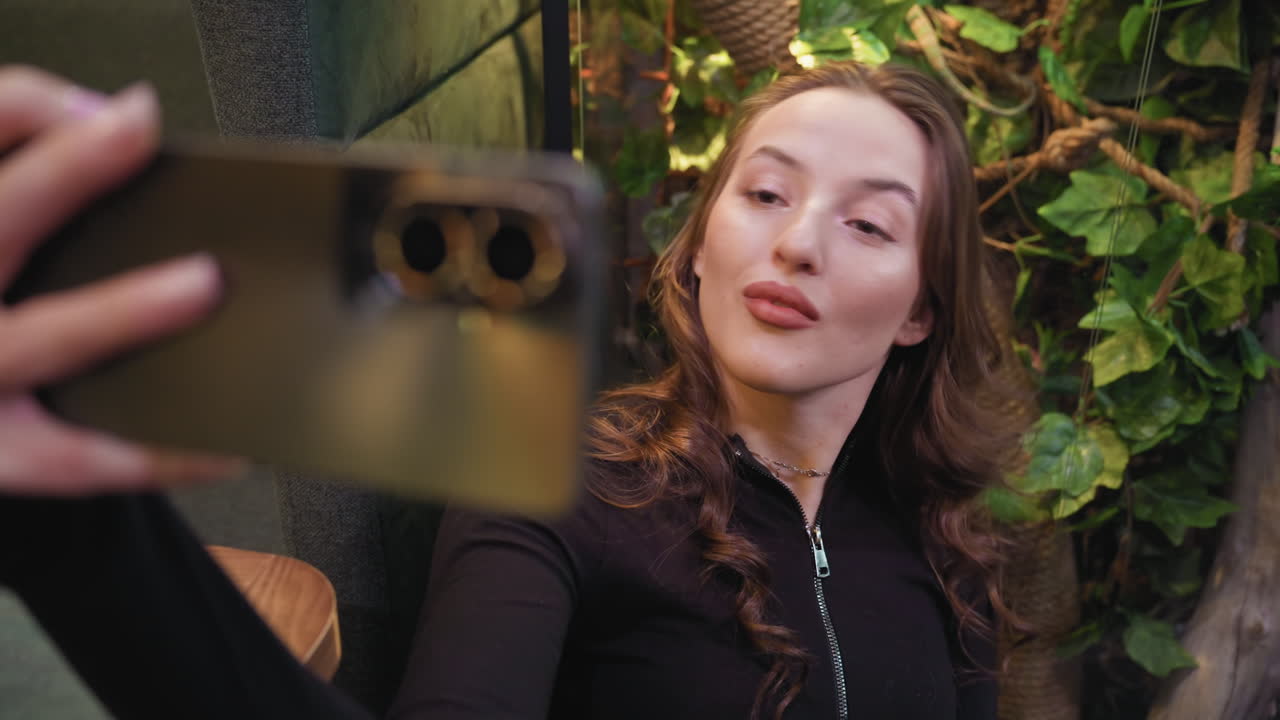 Lady with soft curls poses confidently while taking selfie near lush indoor greenery. Background features decorative foliage, rope textures, and cozy interior atmosphere, with camera phone blurred