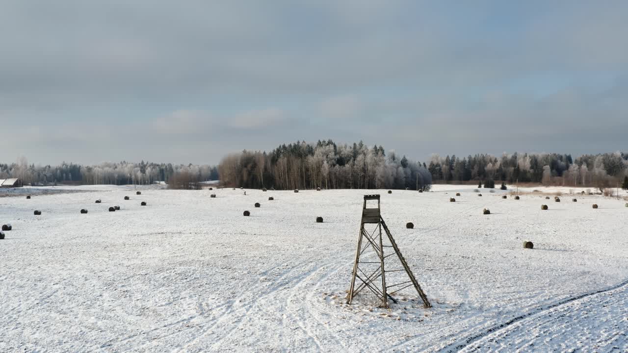 Aerial drone view of wildlife animal hunting tower in a snowy field with hay rolls and a frozen forest in the background on a sunny winter day.
