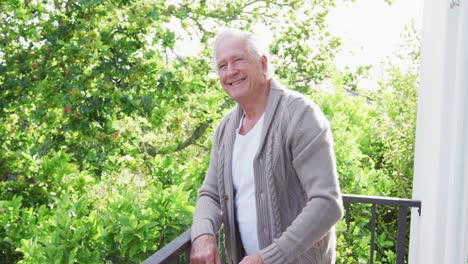 portrait of smiling retired senior man wearing cardigan sweater leaning on railing in balcony