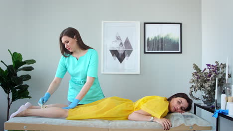 two girls in a beauty salon master in a bathrobe and gloves doing the procedure to remove hair on the legs with a sugar mixture