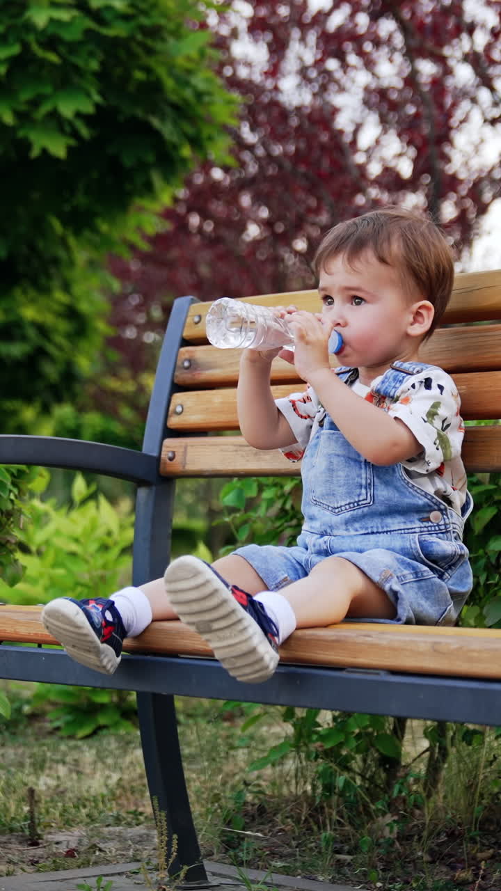 Peaceful little boy sitting on the wooden bench outdoors. Kid drinking water from a bottle. Vertical video
