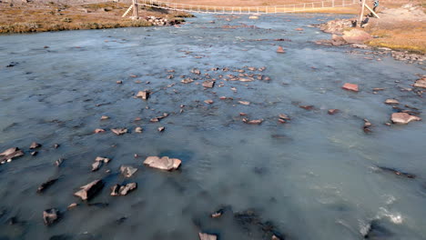 Close-up-tracking-shot-of-a-river,-that-flows-through-a-stony-and-barren-landscape,-a-small-bridge-is-seen,-sunny-day,-daylight,-outdoor,-wilderness