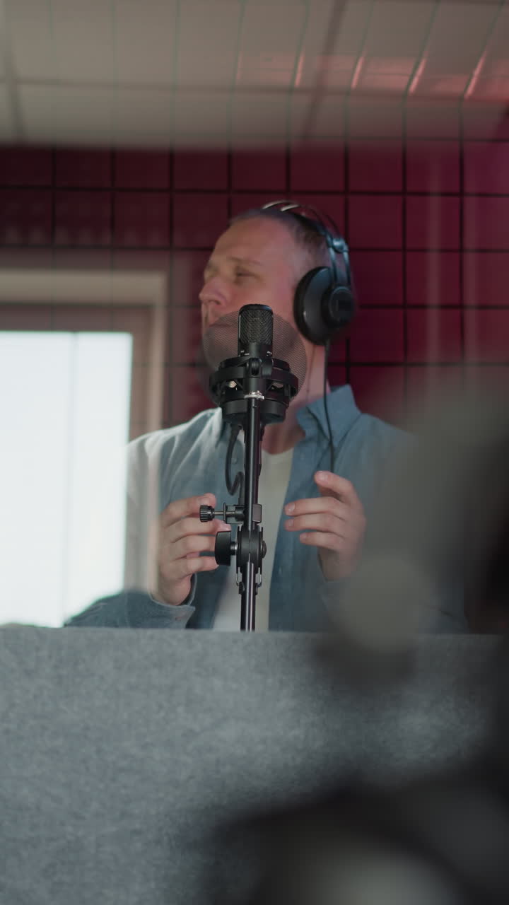 Close-up of a black speaker in a recording studio, with a pair of black headphones. In the background, a man passionately sings while wearing a blue long sleeve shirt. The red acoustic background