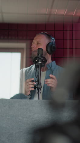 primer plano de un altavoz negro en un estudio de grabación, con un par de auriculares negros. en el fondo, un hombre canta apasionadamente mientras lleva una camisa de manga larga azul. el fondo acústico rojo