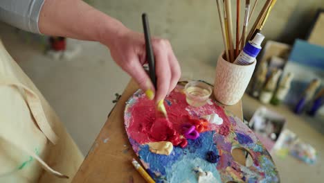 Una-Artista-Femenina-Con-Un-Pincel-Grueso-Está-Mezclando-Pinturas-Al-óleo-De-Diferentes-Colores-En-Una-Paleta:-Morado,-Rojo,-Amarillo.-Vista-Cercana-De-La-Mano-Femenina-Mezclando-Colores.