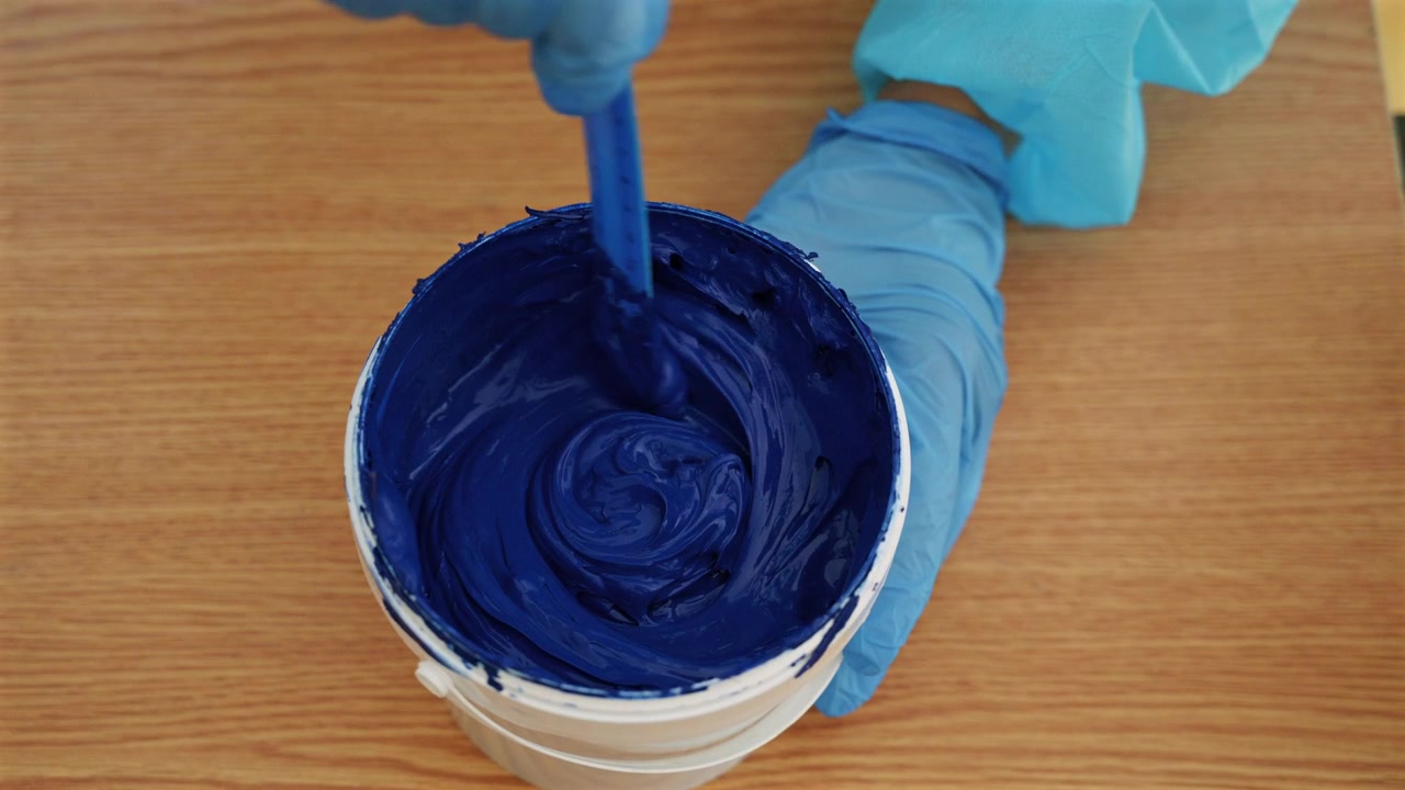 Close-up de tinta azul espessa para serigrafia sendo mexida em um recipiente por um trabalhador com luvas.