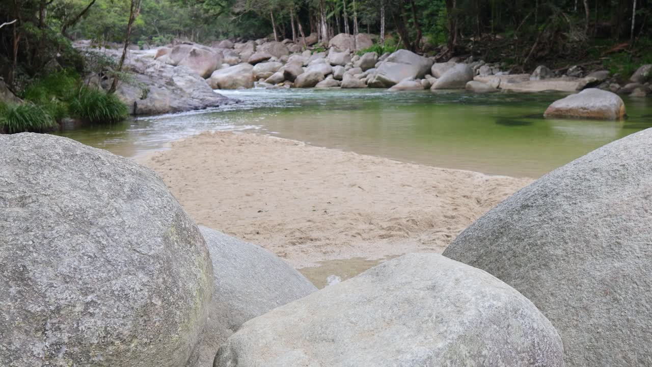 A tranquil creek with clear water flows through lush rainforest, surrounded by large rocks and greenery under soft natural light