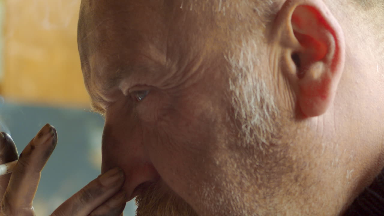 un anciano artista canoso, con la cara sucia de carbón, mira fijamente al frente, fumando un cigarrillo