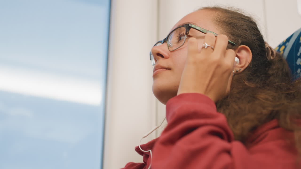 Young woman on train listening earbuds, adjusting glasses, curly hair framing face, looking upward toward sky through window, soft daylight, serene study commute mood, ambient music playing