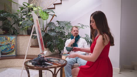 la mujer pone una taza de café en la mesa del artista. el pintor hace una pausa en el proceso artístico evaluando al modelo en vivo en vestido rojo con una mirada contemplativa