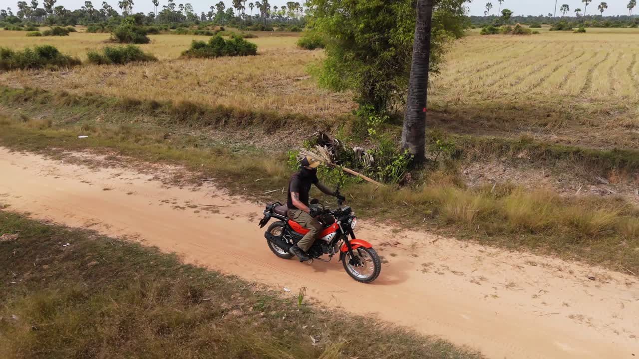 Un drone aereo segue e orbita attorno a un motociclista che percorre una strada sterrata accanto a risaie nella Cambogia rurale