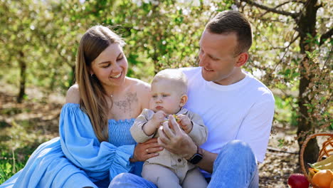parents look with adoration on their lovely little toddler. blond baby sitting in dad's hands looks at the flower. mother kisses her son on the head. nature at backdrop.