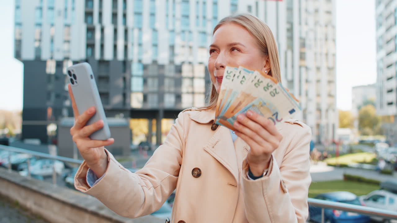 Happy rich mature caucasian businesswoman using smartphone holding euro money cash on city street