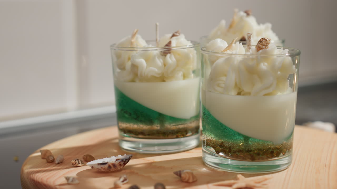 White glass cup filled with layered candle wax in creamy and green tones, topped with elegant whipped wax and tiny shell decorations, creating realistic dessert-like appearance on wooden tray