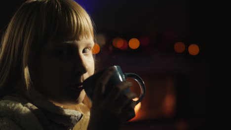 una chica bebe té sentada junto a la ventana, una chimenea y un árbol de navidad están iluminados por detrás