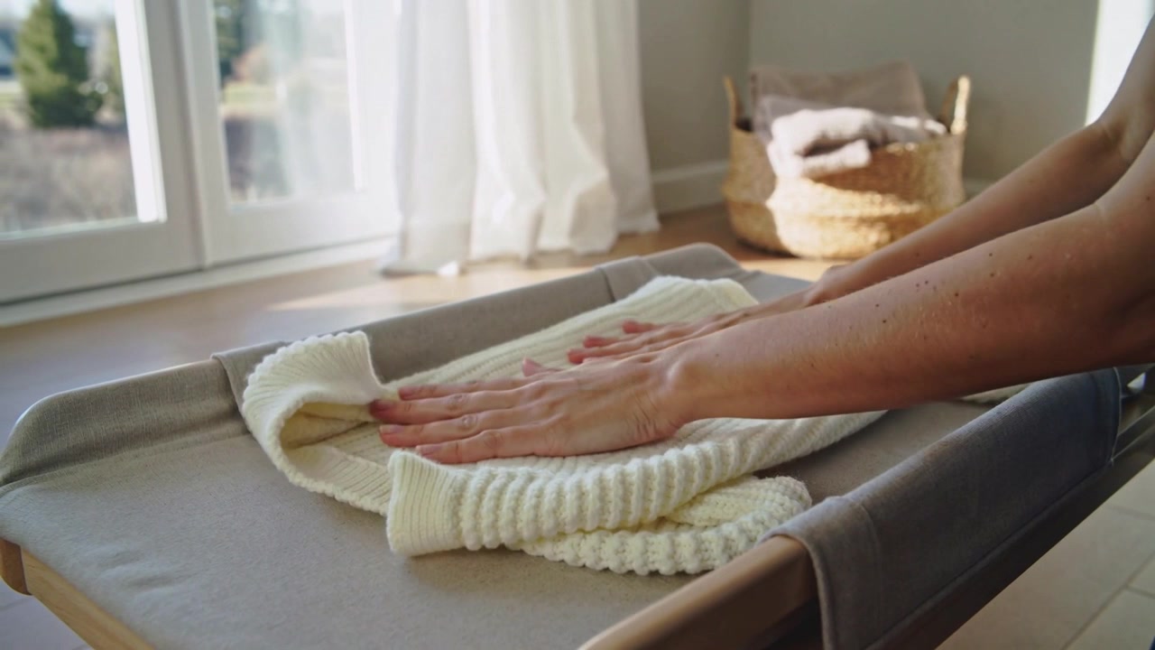 Entering frame, forearms folding and smoothing cream ribbed sweater on pad by window for neatness