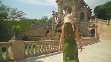 stunning footage of a young caucasian girl in a green dress and hat walks joyfully in front of cascada monumental - gaudí's fountain in parc de la ciutadella