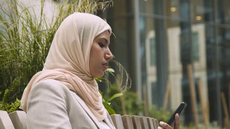 muslim businesswoman answering call on mobile phone sitting outside office in city gardens