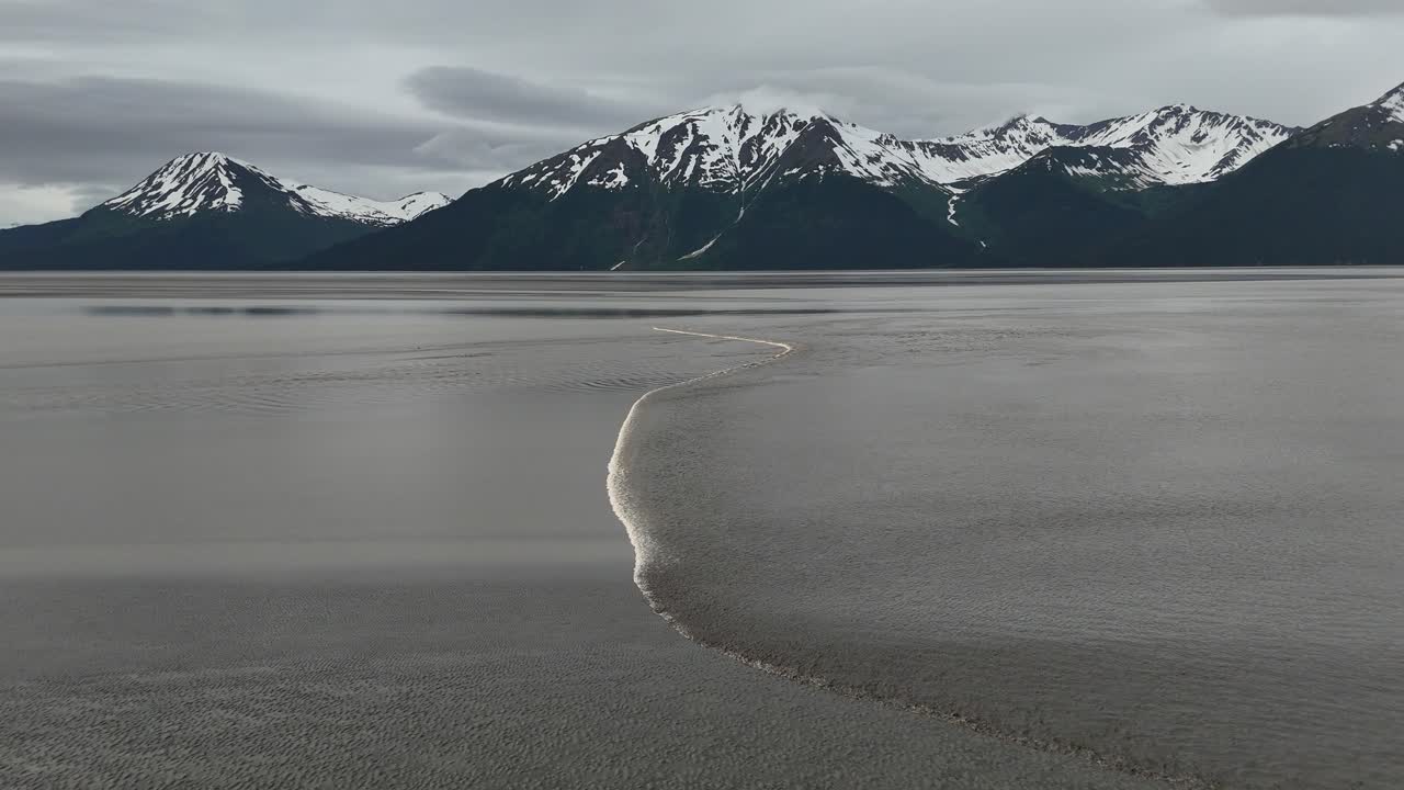 Aerial View of Bore Tide coming in on the Turnagain Arm in Alaska, Small wave of water coming into shore, Mountains in background with patches of snow