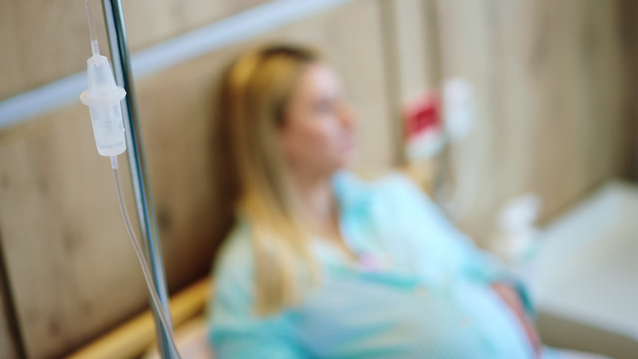 Drop counter connected to a patient lying on bed. Blonde pregnant woman strokes big belly at backdrop in blur.