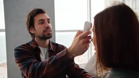 Un-Chico-Moreno-Con-Barba-Incipiente,-Con-Una-Camisa-A-Cuadros-Rojos-Y-Azules-Y-Una-Camiseta-Blanca,-Mide-La-Temperatura-Con-Un-Termómetro-Electrónico-De-Su-Novia-Morena-Con-Un-Suéter-Verde-Mientras-Está-Sentada-En-El-Sofá-De-Un-Moderno-Apartamento-Con-Grandes-Ventanales.