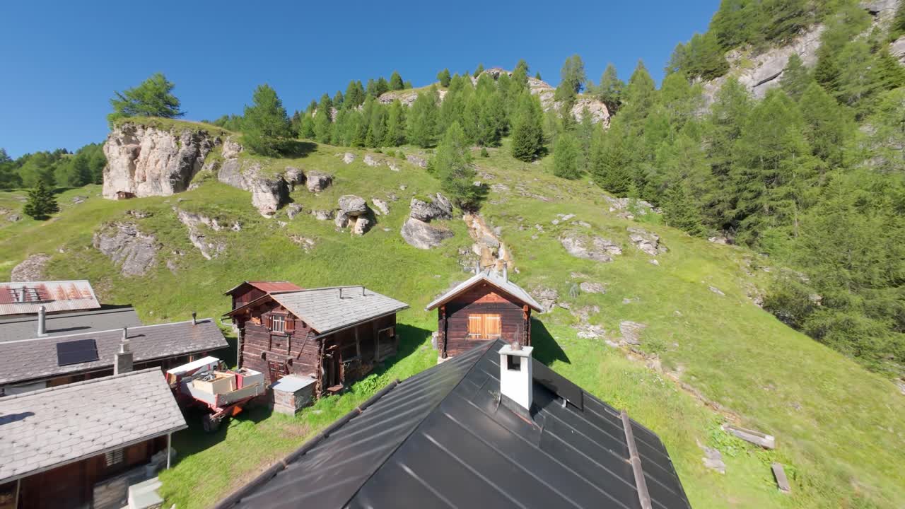 Dron FPV krąży nad Bavugls w pobliżu Andeer w Szwajcarii, ukazując rustykalne alpejskie chaty i drewniane detale. Wyraźny widok na frontowe wejście i balkon pośród iglaków, łąk i górskiej doliny.