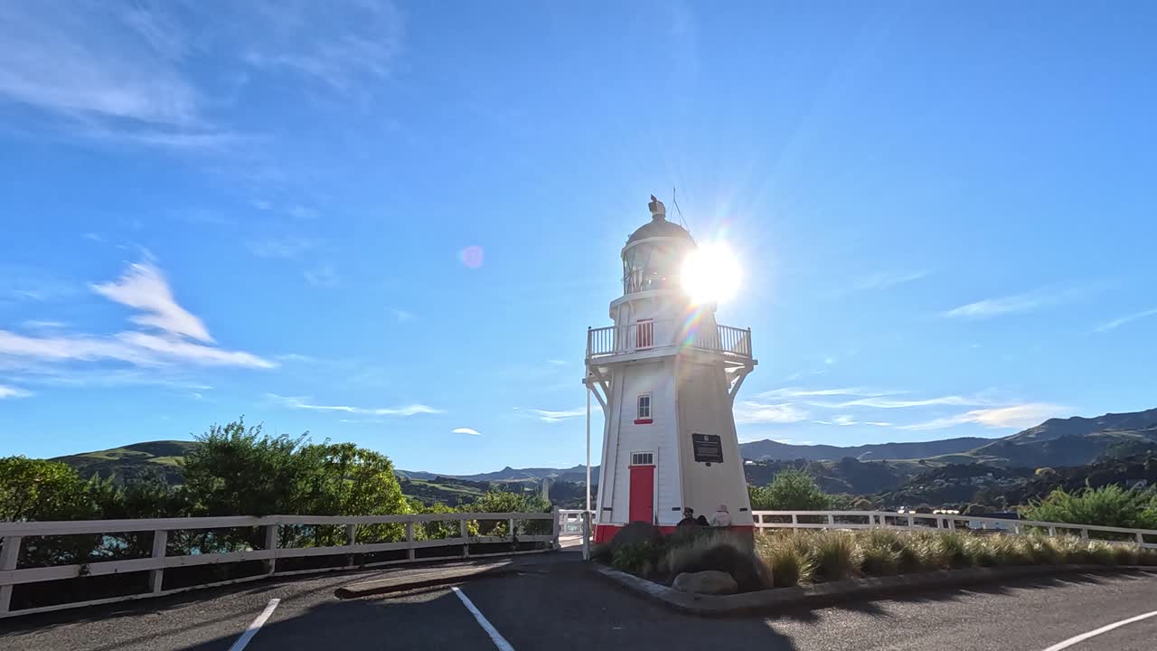 A serene lighthouse stands under bright sunlight, offering panoramic views of Akaroa Harbor and surrounding landscapes
