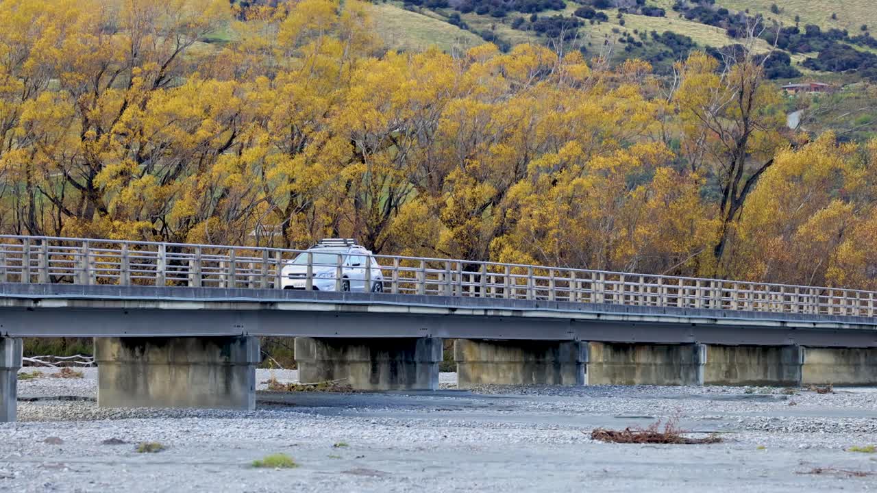 White camper van drives across rural bridge, autumn foliage, daylight, static wide shot, Glenorchy