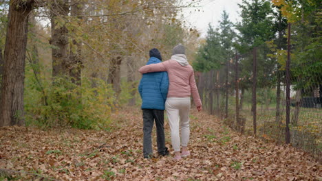 back view of woman in pink top and jogger affectionately holding student in blue sweater as they stroll through autumn leaves in a serene outdoor setting