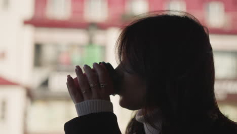 close-up of woman sipping coffee from a cup outdoors, fingers adorned with rings, blurred red building and soft natural light in background creating