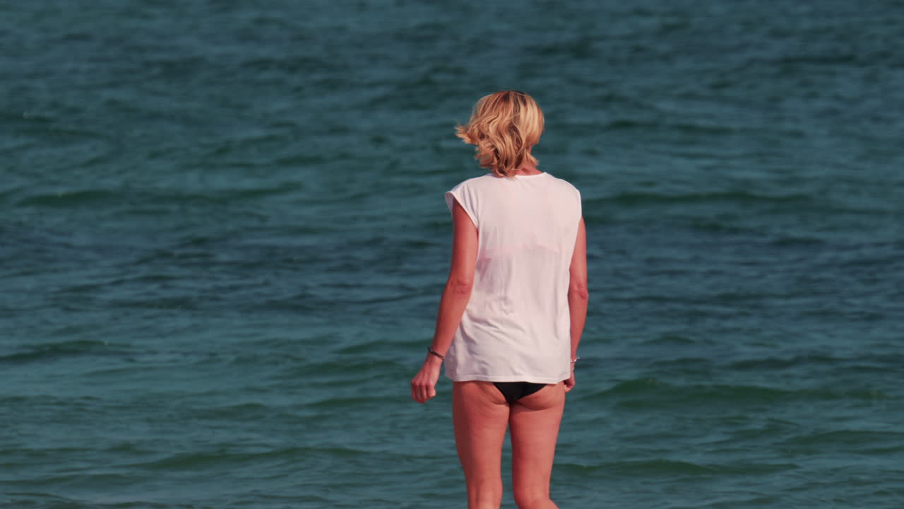 A woman with short blond hair stands waist deep in the sea, facing the horizon