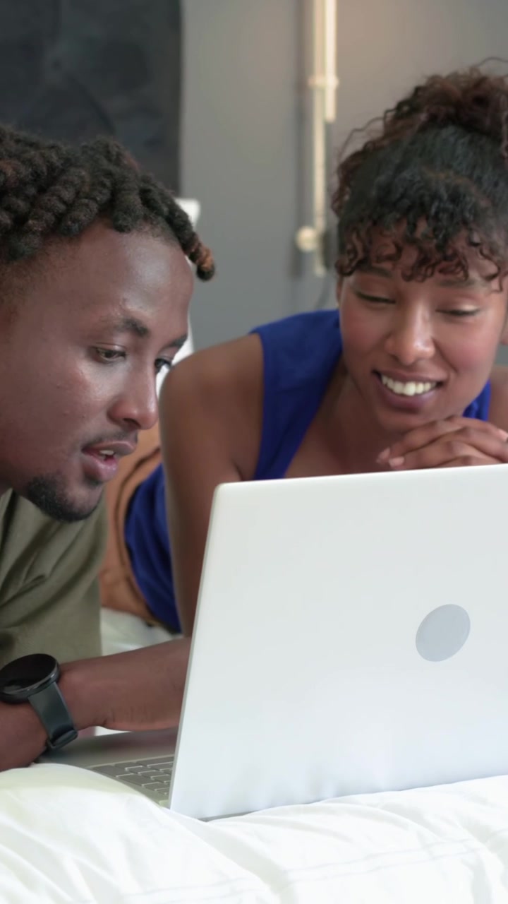 Vertical video: Seeing white laptop, man green shirt leaning talking, woman blue top smiling on bed