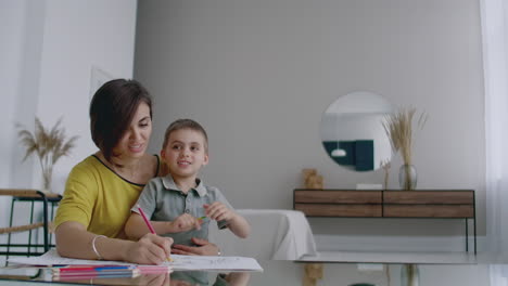 madre e hijo sentados en la mesa del espejo en la habitación blanca dibujando con lápices de colores sonriendo y riendo