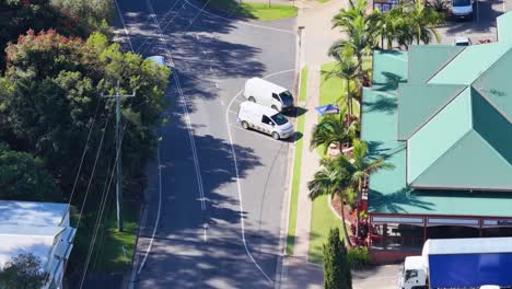 drone captures cars moving past a hotel with green roofs in a sunny, tree-lined street