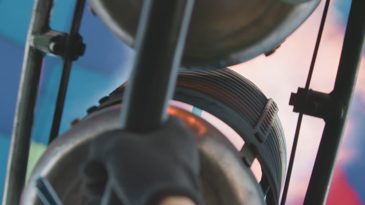 Close up of metal burner flame erupting into vibrant balloon envelope as pilot ignites fuel jets, heat rising in bursts, highlighting coil tubing and frame against colourful fabric background