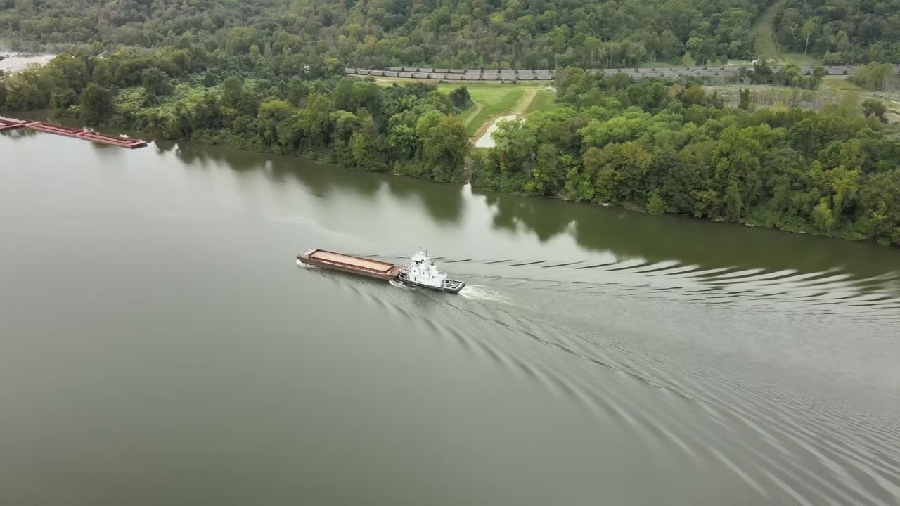 Aerial drone footage of towboat guiding cargo barge along calm Kanawha River surrounded by forested hills, industrial docks, and quiet roads in scenic West Virginia, United States
