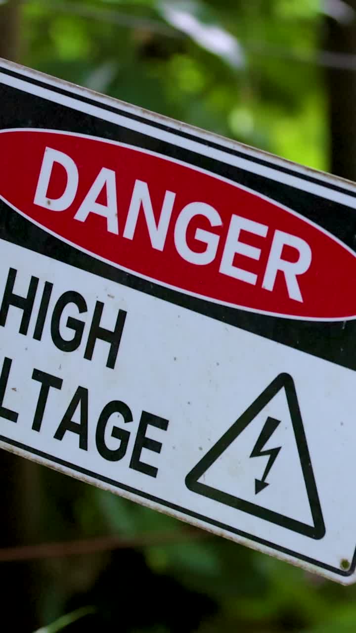 Primer plano vertical de una señal de advertencia de alto voltaje balanceándose contra follaje verde.