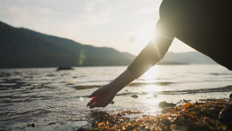 lady gently strokes rippling water of lake under sunlight. woman puts hand down into warm water enjoying soft glow of sunset. connection with rhythms of nature