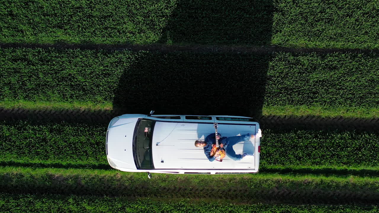 Extreme driving on a roof of a car. Bearded guy lying on a moving car and playing the guitar on green field background. Aerial view.
