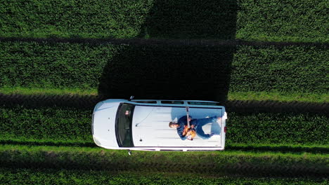 extreme driving on a roof of a car. bearded guy lying on a moving car and playing the guitar on green field background. aerial view.