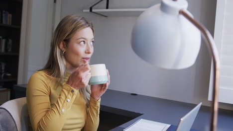 Biracial-woman-sitting-at-desk-using-laptop-and-drinking-coffee-at-home,-slow-motion