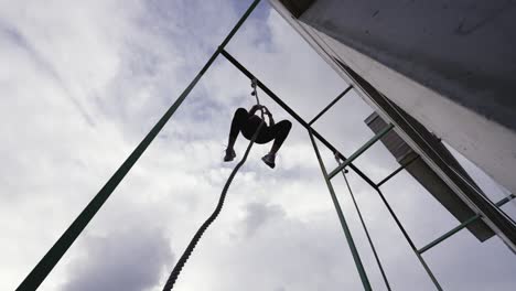 dynamic slow motion footage of a fit woman descending after a successful rope climb, backlit against the clouded sky