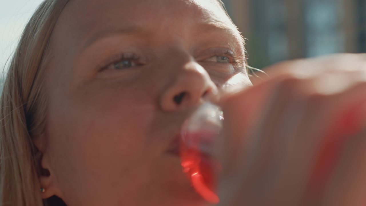 Una donna beve un liquido rosso da una bottiglia di plastica trasparente in primi piani. L'attenzione rimane sul suo viso e sull'azione del bere, con luce naturale soffusa che evidenzia i dettagli della texture della pelle. Sfondo.