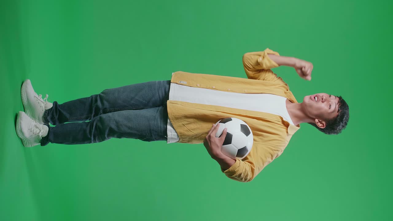 공을 들고 있는 아시아 소년의 몸 전체 측면 뷰, 골을 외치며 우승팀을 축하하면서 초록색 스크린 배경에서 축구를 응원합니다.