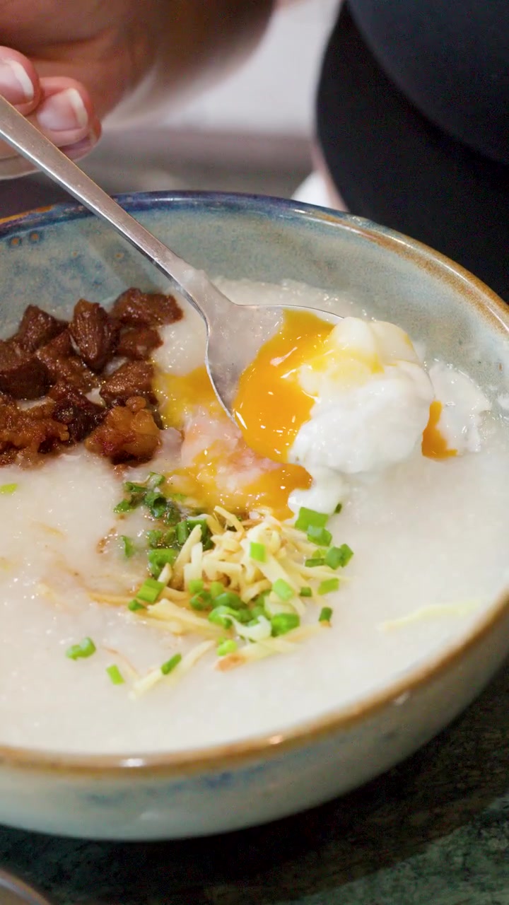 Bidikan jarak dekat sendok memecahkan kuning telur di atas bubur nasi gurih dengan daging babi dan daun bawang