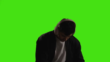 Man-Putting-On-Wireless-Headphones-And-Streaming-Music-From-Mobile-Phone-Against-Green-Screen-With-Low-Key-Lighting